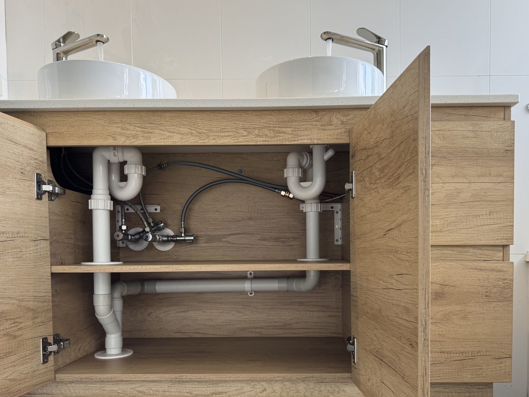 A finished double vanity with the cabinet doors open, showing neatly run and clipped waste and water pipework underneath