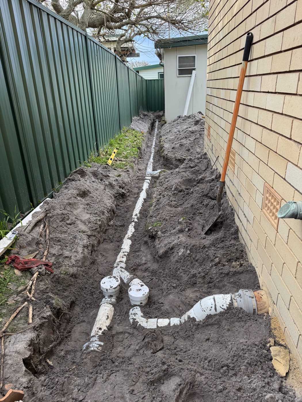 An open trench running alongside a house with new stormwater pipe laid in it, shovel and tape measure in the trench