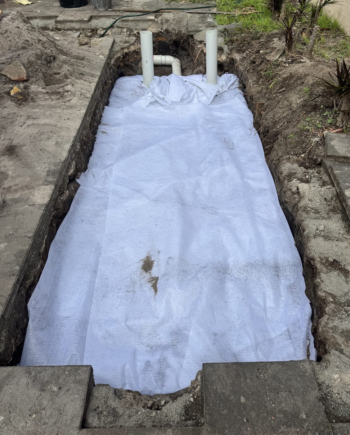 A drainage trench lined with white geotextile fabric wrapped around a pipe run, with two vent stubs visible
