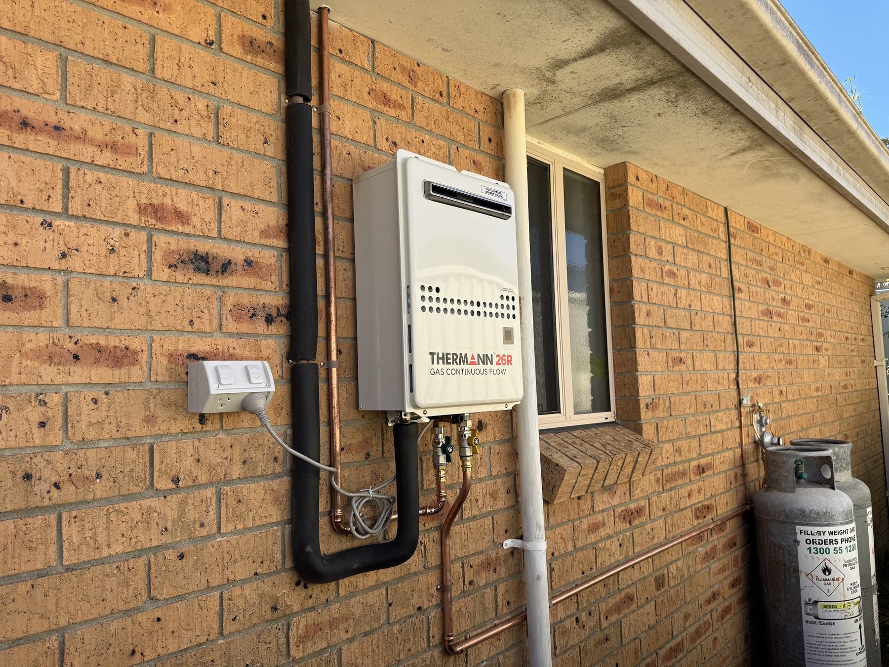 A Thermann continuous flow gas hot water unit installed on a brick wall with neat copper pipework and an LPG cylinder alongside